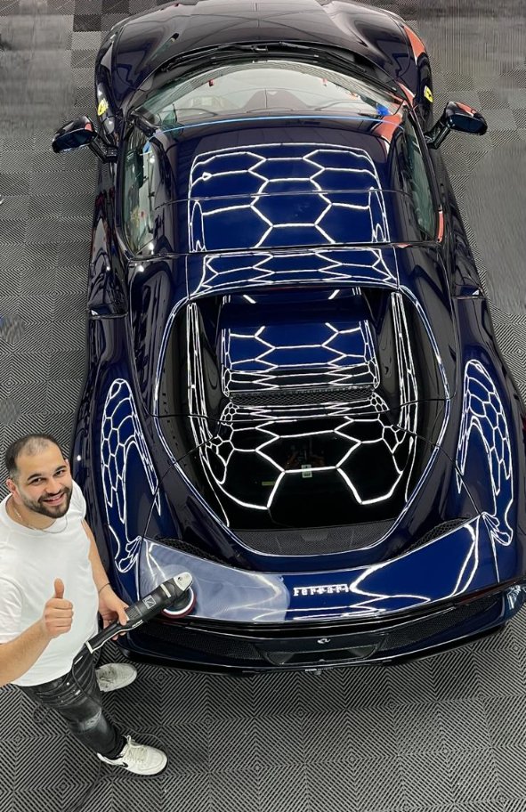 Donkerblauwe Ferrari, van bovenaf gefotografeerd in de cabine van MK Detailing. De hexagon-verlichting van het plafond weerspiegelt in het lak. Links onderin staat de eigenaar met een polijstmachine.