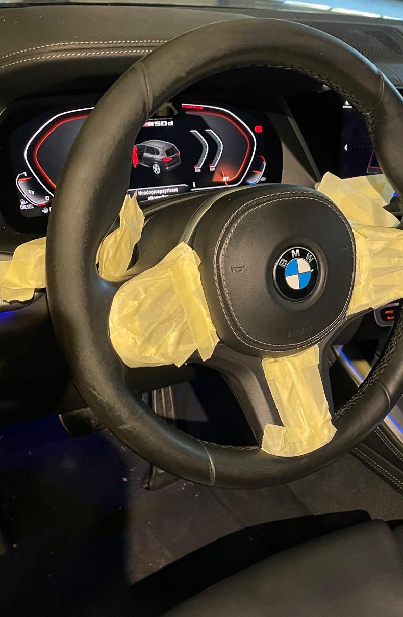 Stuurwiel van een BMW met de spaken en de airbag afgeplakt met gele tape voordat er aan het interieur wordt gewerkt.