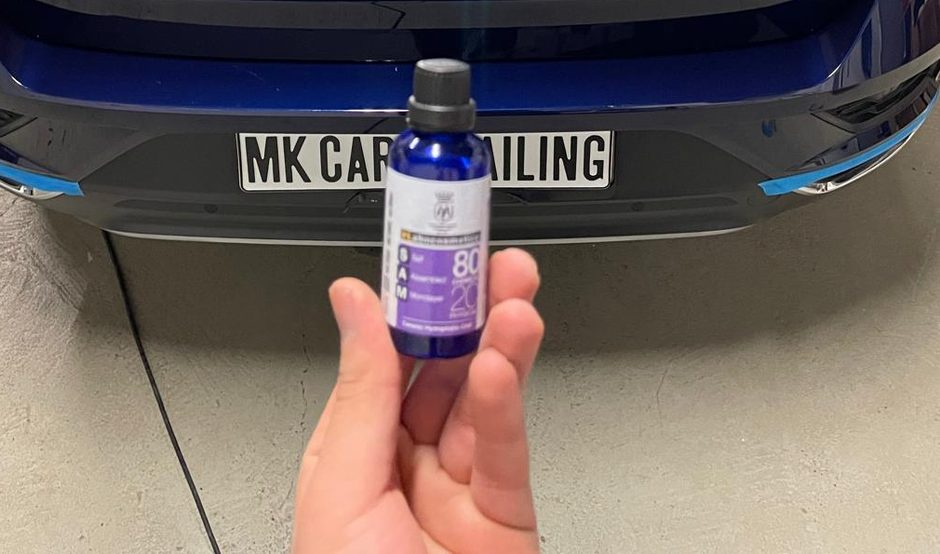 Hand met een flesje keramische coating voor de achterbumper van een auto waarvan de randen zijn afgeplakt met blauwe tape.
